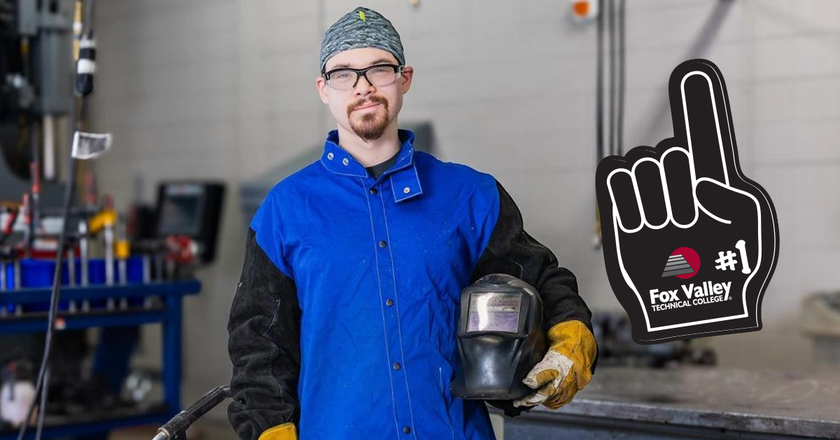 student smiling at camera and holding a welding helmet in a shop with Fox Valley Tech Number 1 sticker