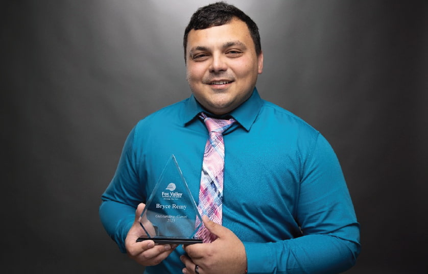 Image of man in blue shirt and pink tie holding a glass, triangular shaped award with Bryce Remy Outstanding alumni award engraved on it. 