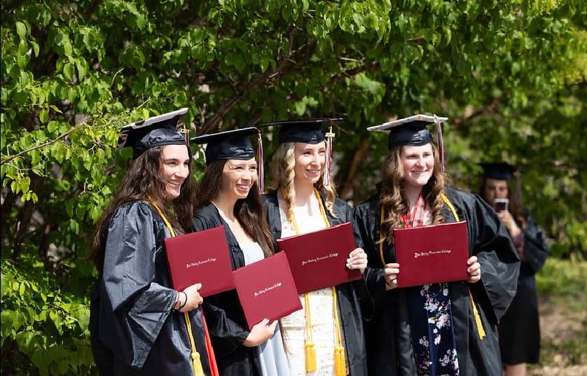 Four graduates in caps and gowns stand outdoors, smiling and holding red diploma covers. Trees with green leaves are in the background. Another person is visible behind them, taking a photo.